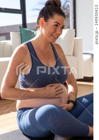 Pregnant woman sitting cross-legged on yoga mat in living room, cradling belly and smiling Pregnant woman sitting cross-legged on yoga mat in living room, cradling belly and smiling 140930413