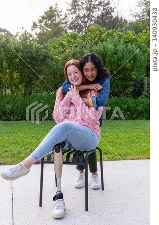 Diverse women embracing on metal chair on concrete patio in garden with prosthetic leg, sneakers 140930438
