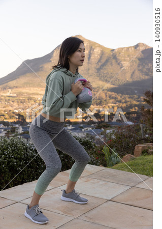 Asian korean woman performing weighted squat in sportswear on tiled terrace holding kettlebell Asian korean woman performing weighted squat in sportswear on tiled terrace holding kettlebell 140930516