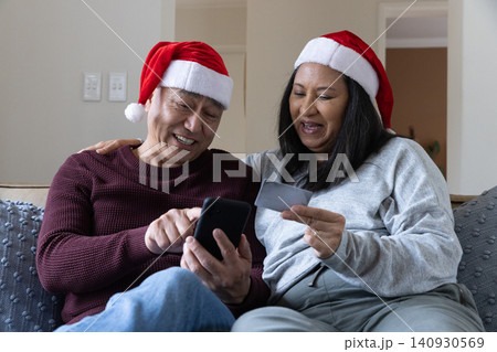 Senior couple in santa hats holding smartphone and credit card leaning on sofa in living room Senior couple in santa hats holding smartphone and credit card leaning on sofa in living room 140930569