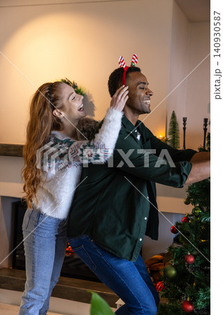 Diverse couple decorating christmas tree in living room with candy cane, headband and lit fireplace Diverse couple decorating christmas tree in living room with candy cane, headband and lit fireplace 140930587
