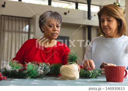 Diverse senior and middle-aged women crafting holiday wreath at home with fir sprigs pinecones 140930613