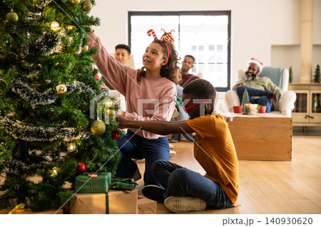 African american children kneeling by home christmas tree hanging ornaments, tinsel, copy space African american children kneeling by home christmas tree hanging ornaments, tinsel, copy space 140930620