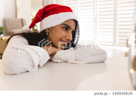 Woman leaning on tabletop in living room wearing knit sweater with Santa hat near Christmas tree Woman leaning on tabletop in living room wearing knit sweater with Santa hat near Christmas tree 140930626
