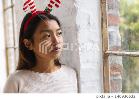 Asian woman leaning by loft window wearing candy-cane antler headband facing raindrops, copy space Asian woman leaning by loft window wearing candy-cane antler headband facing raindrops, copy space 140930632