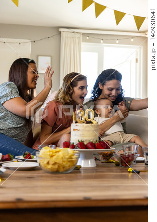 Diverse female friends with infant posing for birthday selfie on couch in living room with cake Diverse female friends with infant posing for birthday selfie on couch in living room with cake 140930641