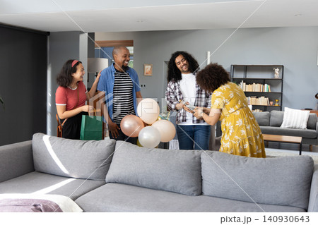Diverse friends gathering in living room at sofa, handing wrapped gift, holding pastel balloons Diverse friends gathering in living room at sofa, handing wrapped gift, holding pastel balloons 140930643