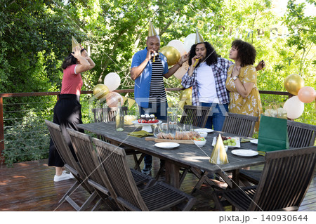 Diverse friends wearing party hats blowing noisemakers around table on deck with cake, balloons 140930644