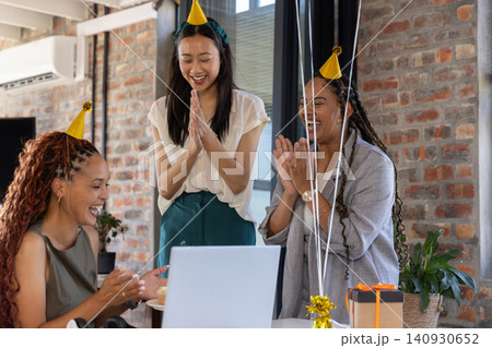 Diverse female coworkers in party hats clapping and smiling around laptop with gift box at office 140930652