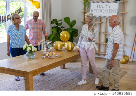 Diverse senior friends standing around wooden dining table in living room admiring balloons 140930685