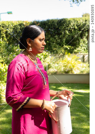 Indian woman wearing pink kurta and watering plants with watering can in sunny backyard, copy space 140930895