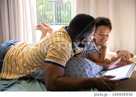 African American father and daughter lying in softly lit bedroom sharing tablet beside teddy bear African American father and daughter lying in softly lit bedroom sharing tablet beside teddy bear 140930936