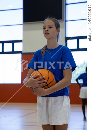 Teenage female basketball player holding basketball standing on gym floor with painted lines Teenage female basketball player holding basketball standing on gym floor with painted lines 140931019
