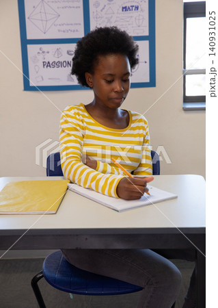 African american female teen writing in notebook with pencil at desk in classroom African american female teen writing in notebook with pencil at desk in classroom 140931025