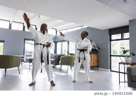 Diverse senior couple performing mirrored karate blocks in living room wearing gis and belts Diverse senior couple performing mirrored karate blocks in living room wearing gis and belts 140931086