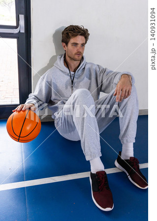 Man wearing tracksuit sitting on court near window and door frame holding orange basketball 140931094