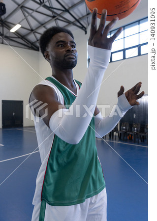 African american man in jersey holding orange basketball overhead in gym with chairs on court 140931095