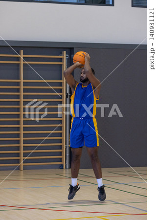 African american male basketball player jumping, shooting orange ball in gym with stall bars African american male basketball player jumping, shooting orange ball in gym with stall bars 140931121
