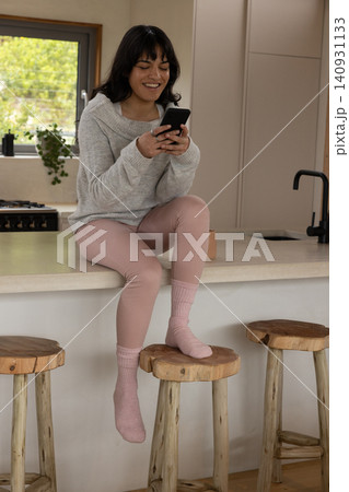 Woman sitting on kitchen island scrolling smartphone beside stove, potted plant on window sill 140931133