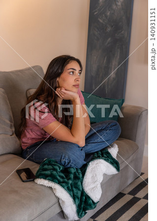 Woman sitting cross-legged on couch in living room gazing right with smartphone and fleece blanket Woman sitting cross-legged on couch in living room gazing right with smartphone and fleece blanket 140931151