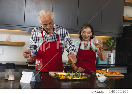 Senior couple pouring oil over vegetables in glass dish at kitchen counter while holding wine glass Senior couple pouring oil over vegetables in glass dish at kitchen counter while holding wine glass 140931163