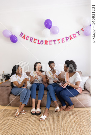 Diverse female friends wearing sashes toasting champagne flutes at home for bachelorette party Diverse female friends wearing sashes toasting champagne flutes at home for bachelorette party 140931231