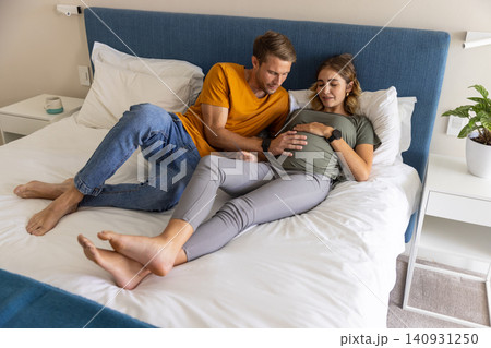 Couple lying on bed touching pregnant belly and sharing glances near nightstands with plant 140931250