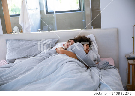 Two men cuddling on bed under light gray duvet in master bedroom near smartphone on nightstand Two men cuddling on bed under light gray duvet in master bedroom near smartphone on nightstand 140931279