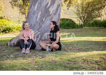 Diverse female friends in activewear sitting by tree in park holding smartphone, water bottle 140931286