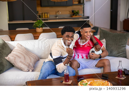 Diverse male friends celebrating and watching match on sofa in living room with pizza, soccer ball 140931296