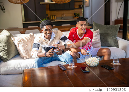 Diverse male friends holding game controllers and grabbing popcorn from coffee table in living room 140931298