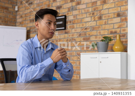 Asian man sitting at wooden table in office with exposed brick wall, wearing light blue shirt Asian man sitting at wooden table in office with exposed brick wall, wearing light blue shirt 140931355