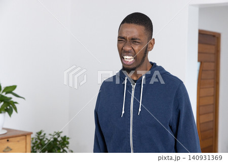 Adult African American man grimacing with eyes closed, standing in room in navy hoodie near dresser Adult African American man grimacing with eyes closed, standing in room in navy hoodie near dresser 140931369