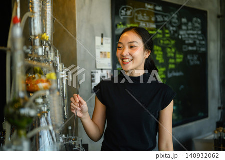 Smiling Young Asian Woman Operating Craft Distillery Equipment in Modern Artisan Workshop Smiling Young Asian Woman Operating Craft Distillery Equipment in Modern Artisan Workshop 140932062