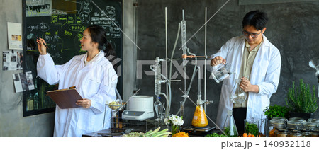 Two Asian Scientists in Lab Coats Conducting Herbal Distillation Experiment with Fresh Botanicals and Glass Apparatus 140932118