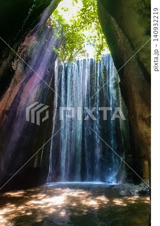Cave waterfall viewed from inside rocky grotto with light rays filtering through jungle canopy opening above 140932219