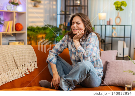 Young woman on home sofa suffering tooth pain after drinking water and feeling worse at home alone 140932954