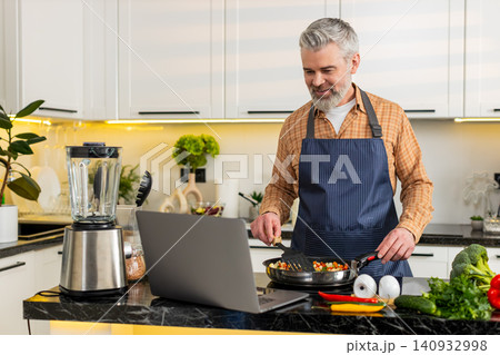 Middle-aged man cooks seasoned vegetables on electric stove watching laptop recipe for healthy diet Middle-aged man cooks seasoned vegetables on electric stove watching laptop recipe for healthy diet 140932998