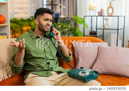 Indian man on home sofa listening to old telephone call with surprised worried face at home indoors Indian man on home sofa listening to old telephone call with surprised worried face at home indoors 140933059