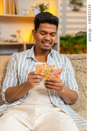 Indian man on home sofa counting cash money in living room with satisfied smile and personal success 140933134