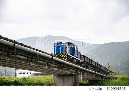 曇り空の下で緑豊かな山並みを背景に高架橋の上を走る青い貨物列 曇り空の下で緑豊かな山並みを背景に高架橋の上を走る青い貨物列 140934208