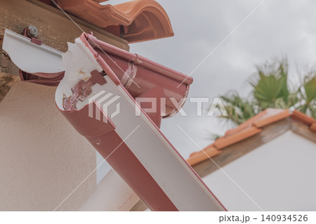 Close up shot of a broken and disconnected plastic rain gutter on a tiled residential house roof edge needing repair work under a cloudy sky Close up shot of a broken and disconnected plastic rain gutter on a tiled residential house roof edge needing repair work under a cloudy sky 140934526