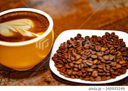 Cup of coffee decorated with a heart and coffee beans on a white plate. dark background Cup of coffee decorated with a heart and coffee beans on a white plate. dark background 140935246