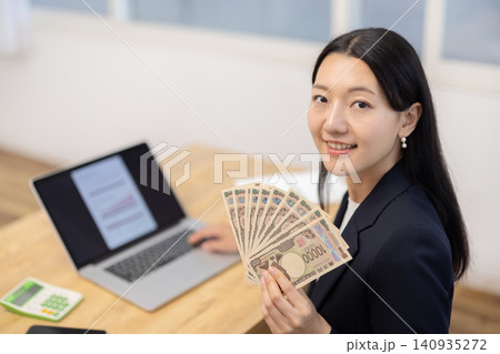 デスクで10万円を手にカメラ目線で微笑む若い日本人の女性 140935272
