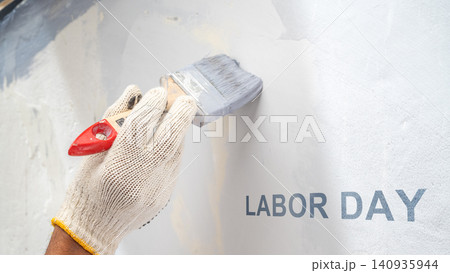 A worker's gloved hand holding a paintbrush, painting gray paint on a wall with the text 'Labor Day' printed on it, representing holiday celebration and manual labor A worker's gloved hand holding a paintbrush, painting gray paint on a wall with the text 'Labor Day' printed on it, representing holiday celebration and manual labor 140935944