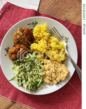 A plate holds turmeric rice, fresh salad, hummus, and two carrot and zucchini meatballs. This meal offers a healthy option for breakfast or lunch enjoyed at the dining table A plate holds turmeric rice, fresh salad, hummus, and two carrot and zucchini meatballs. This meal offers a healthy option for breakfast or lunch enjoyed at the dining table 140936108