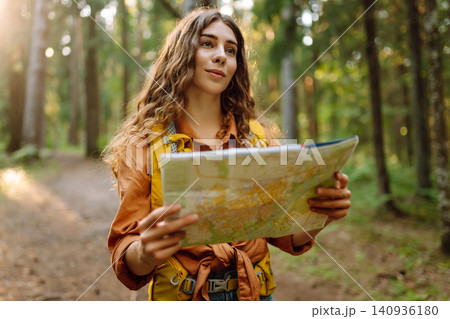 A traveler woman stands in a lush forest, examining a map with a focused expression A traveler woman stands in a lush forest, examining a map with a focused expression 140936180