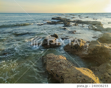 Golden hour light illuminates a rugged path of rocks leading into the calm ocean. This serene seascape symbolizes strength and tranquility, ideal for travel, wellness, and nature projects. 140936233