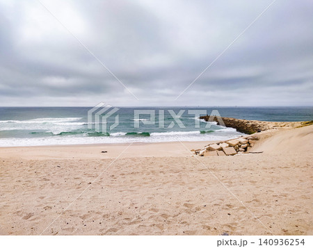 A tranquil, empty sandy beach meets the churning turquoise ocean under a moody, overcast sky. A stone breakwater stretches into the waves, evoking peace, solitude, and natural drama. A tranquil, empty sandy beach meets the churning turquoise ocean under a moody, overcast sky. A stone breakwater stretches into the waves, evoking peace, solitude, and natural drama. 140936254