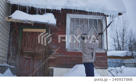 Man removing dangerous icicles and snow from rustic wooden house roof in winter 140937866